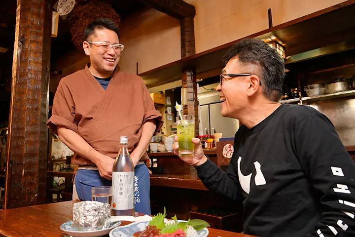 談笑する「居酒屋よさく」の店主・伊藤秀幸さんと丸尾さん